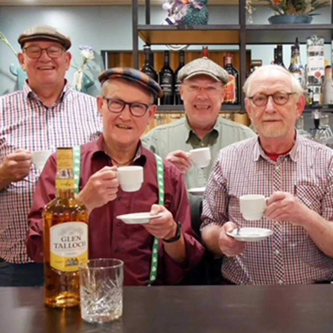 Four older men wearing hats and glasses smiling while holding small white coffee cups and saucers.