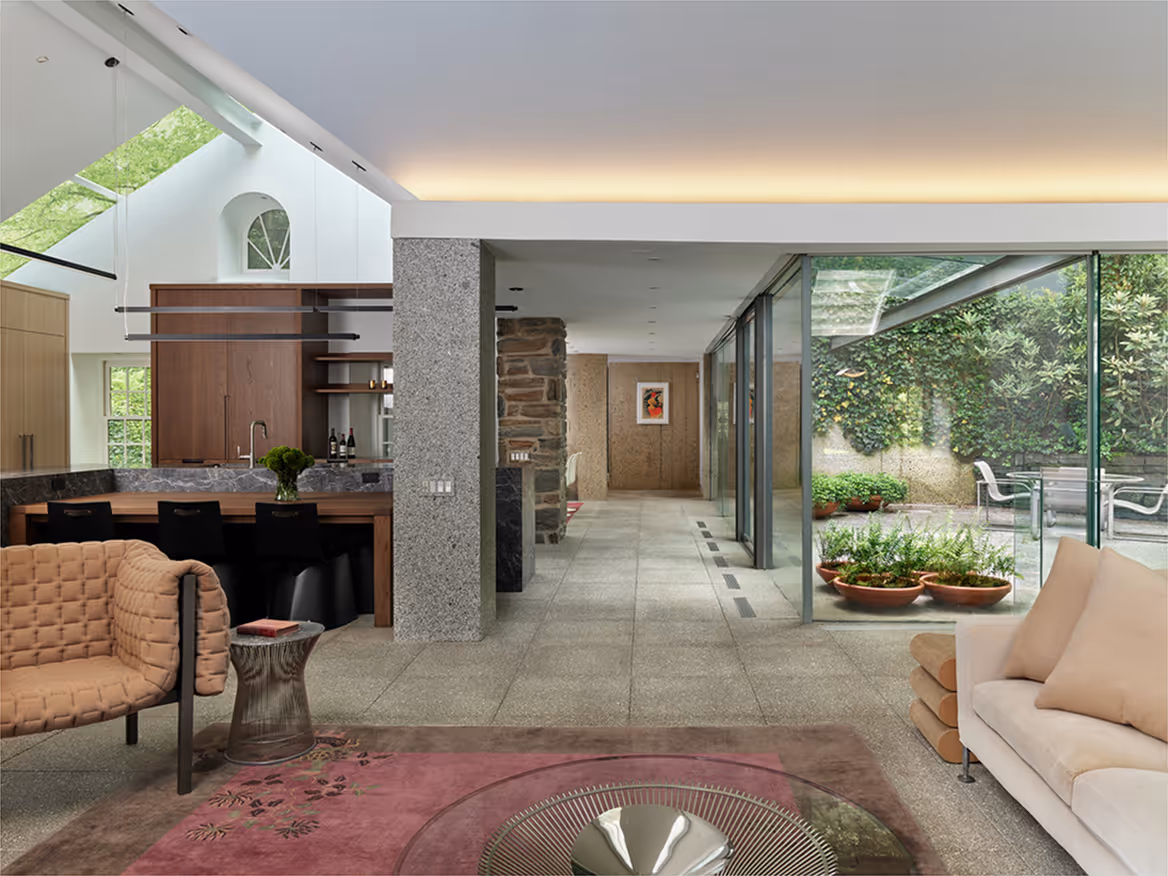 Modern living room and kitchen area with large glass walls overlooking a green outdoor patio.
