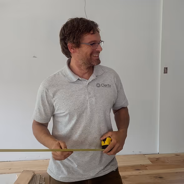 Man wearing glasses and a gray Clarity polo shirt holding a yellow tape measure in a room with unfinished wooden flooring.
