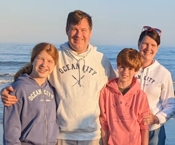Smiling family of four wearing Ocean City hoodies standing by the ocean shore at sunset.