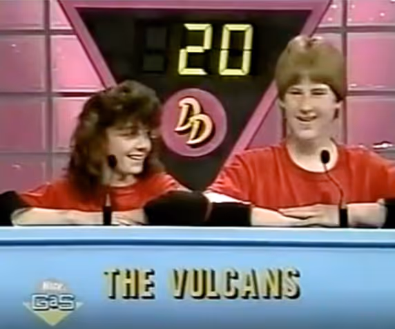 Two teenagers in red shirts smiling behind a game show podium labeled 'THE VULCANS' with a digital score of 20 above them.