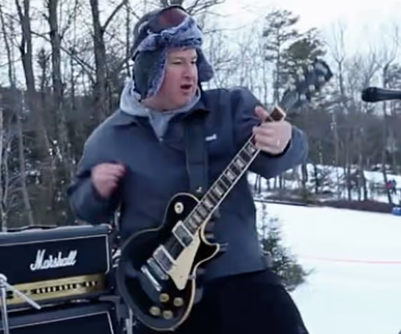 Man wearing a winter hat and jacket playing an electric guitar outdoors in a snowy forest.