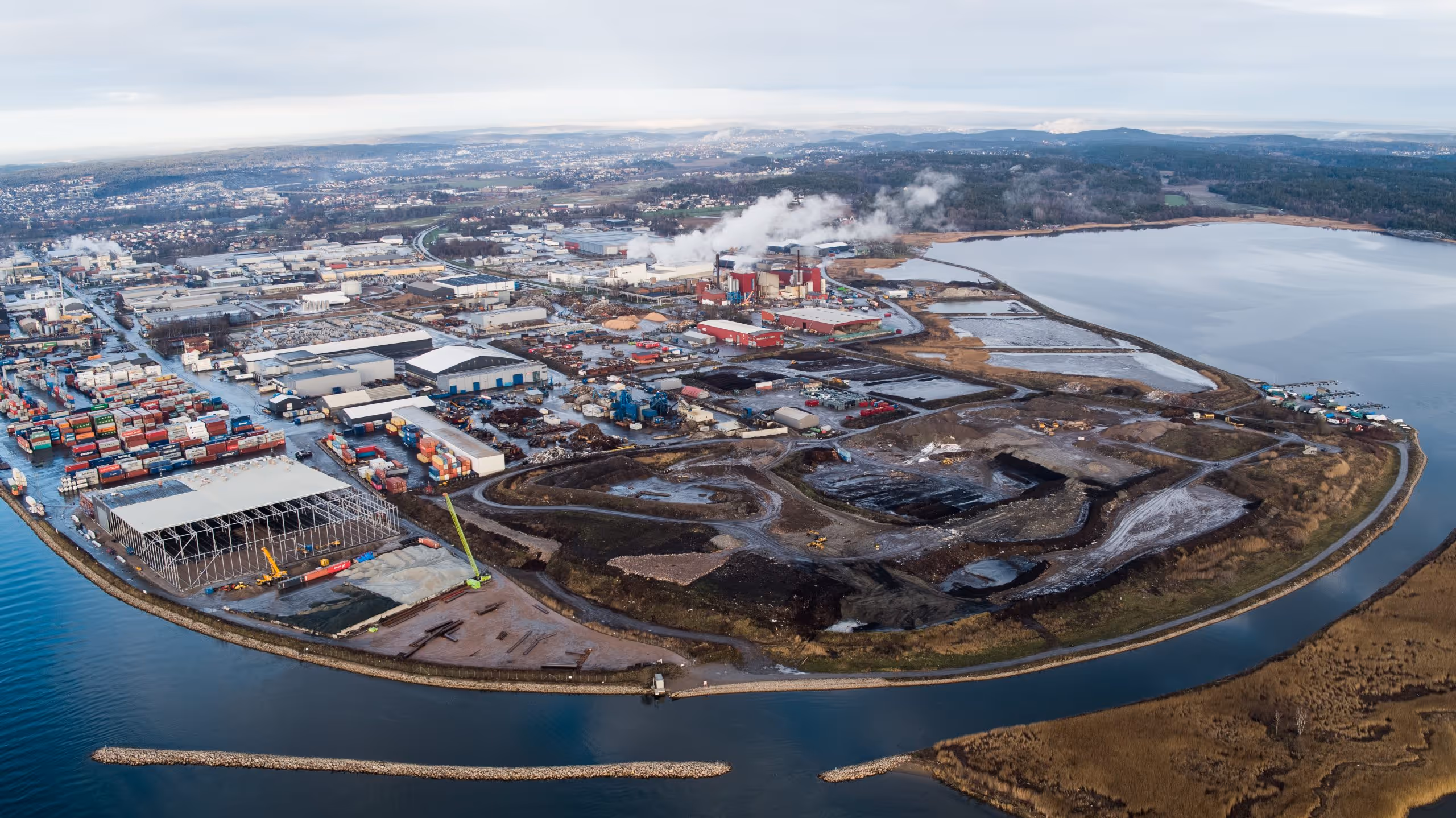 Aerial view of an industrial area with warehouses, shipping containers, smokestacks, and surrounding water.