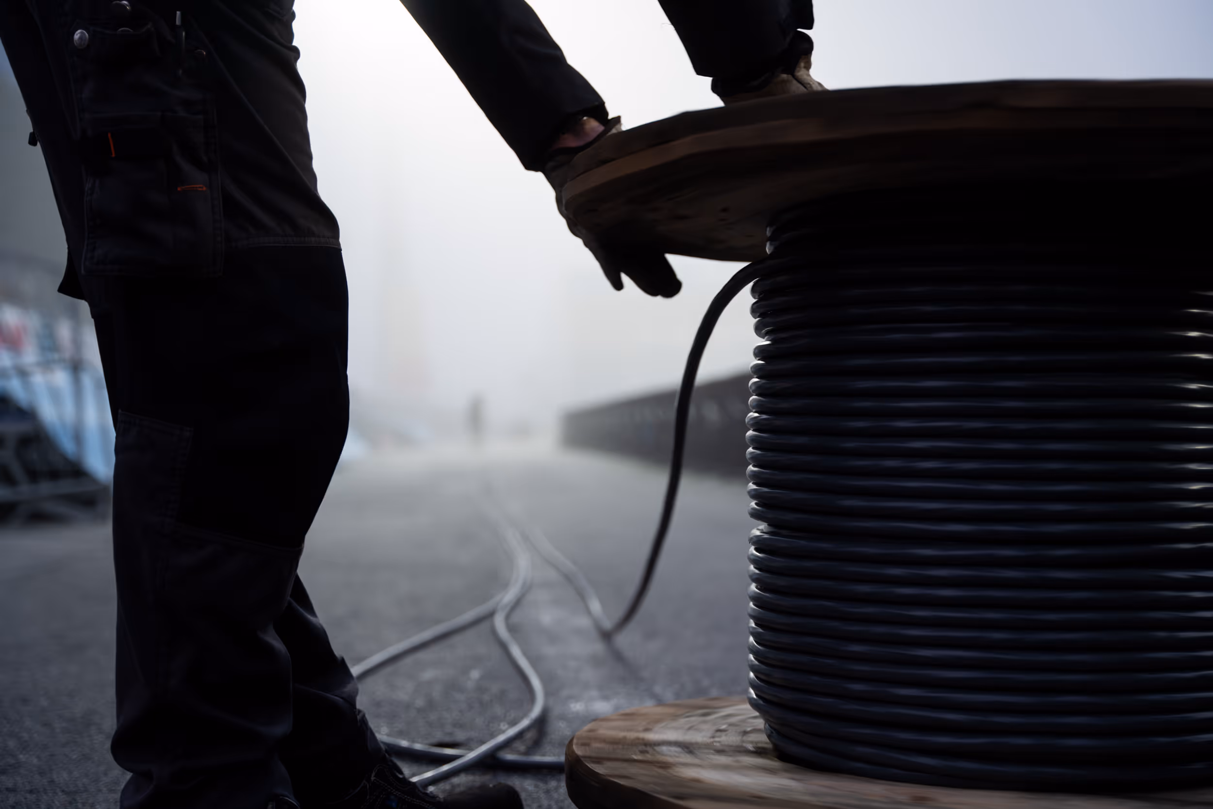 Worker handling a large spool of thick black cable on an outdoor paved surface in foggy weather.