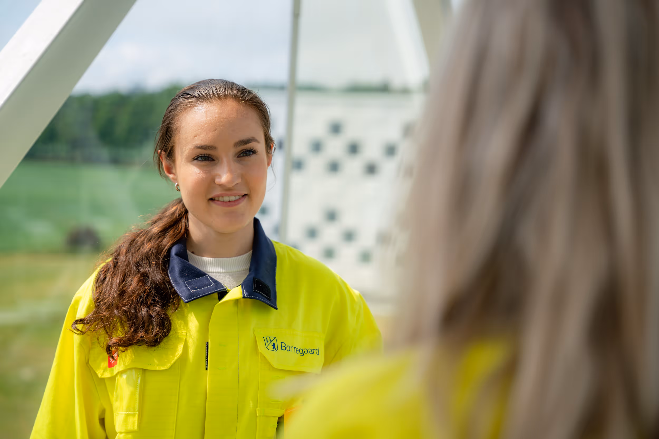 Smilende kvinne i gul arbeidsjakke snakker med en kollega utendørs.