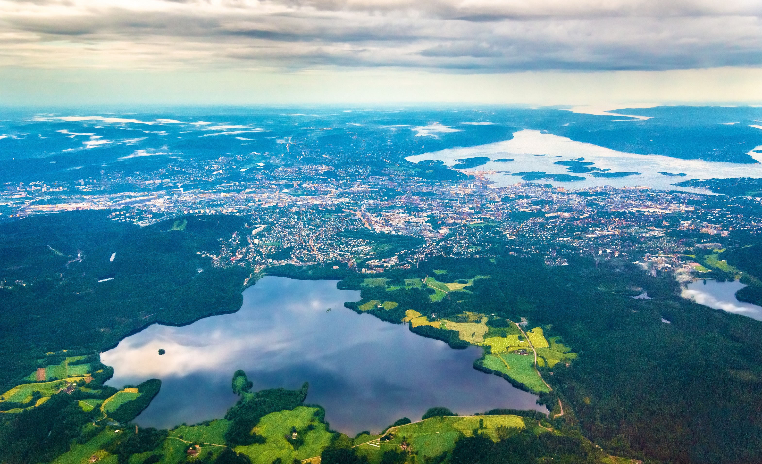 Aerial view of a city by a large lake with surrounding green fields and forests under a cloudy sky.