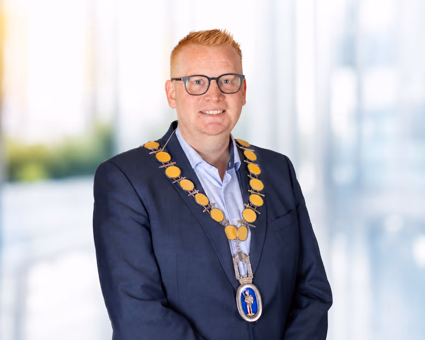 Man with glasses wearing a dark suit and ceremonial gold chain necklace on blurred background.
