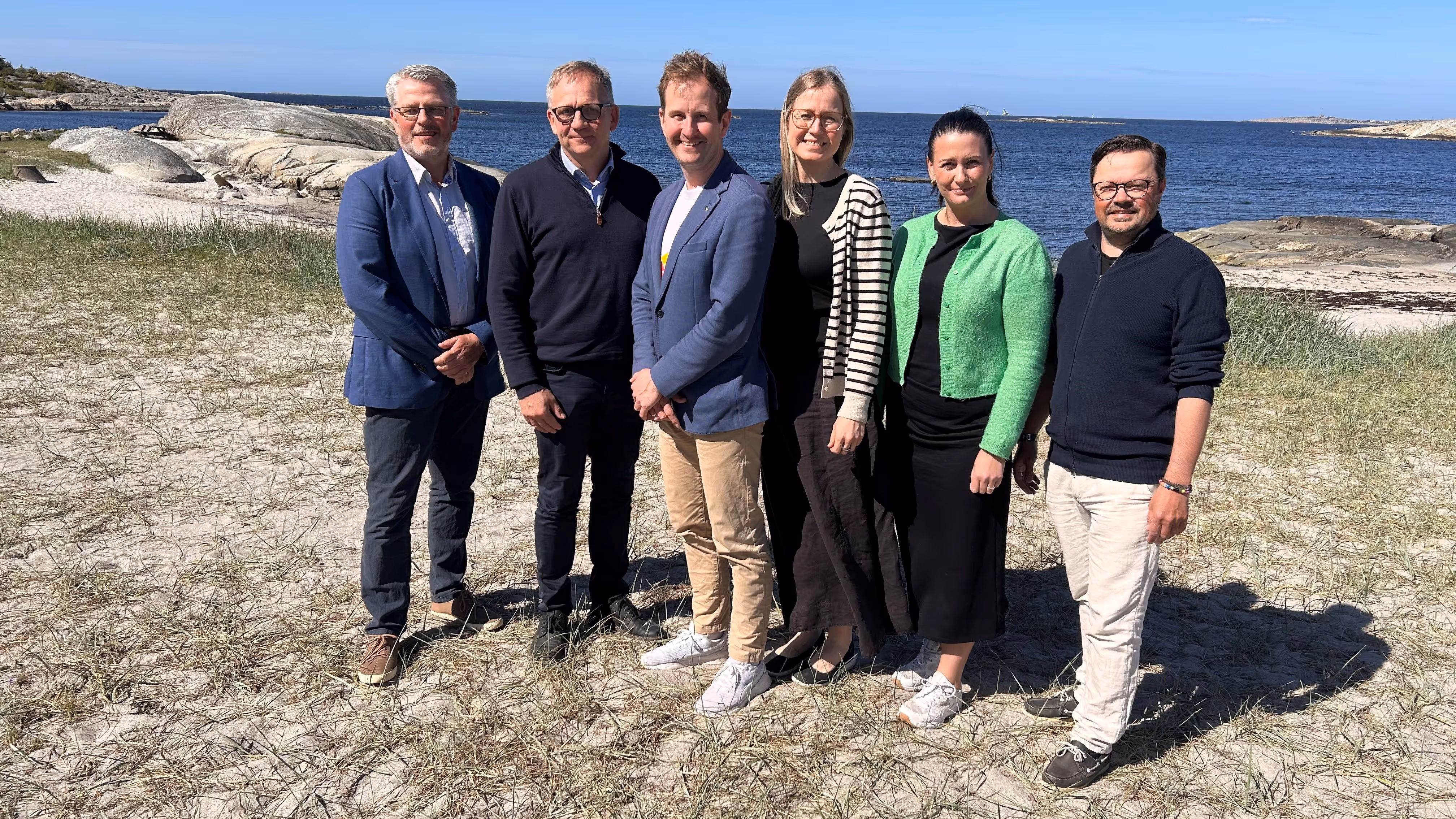 Six people standing on a beach with grassy sand and rocky shore under a clear blue sky.