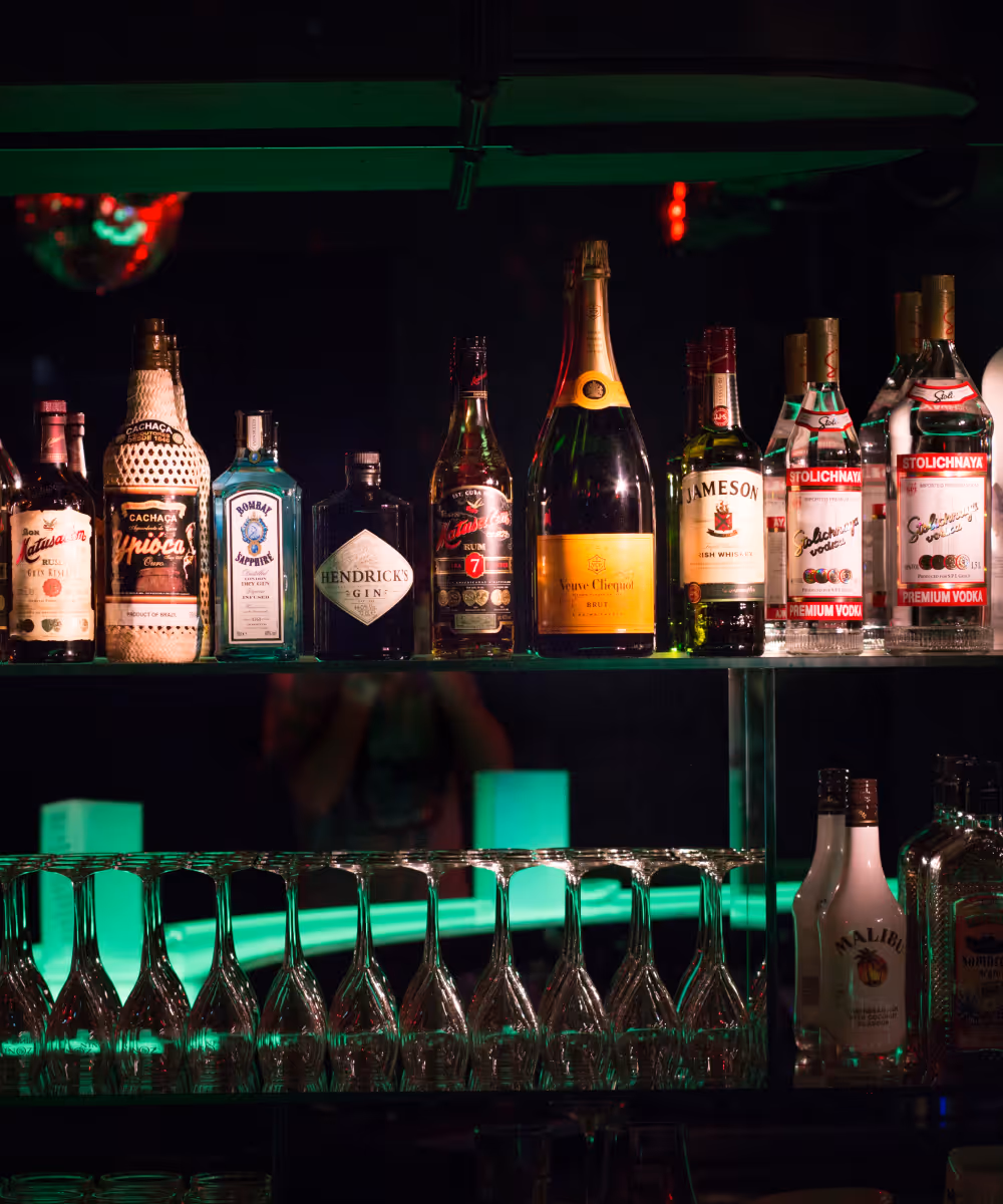 Verschiedene Flaschen alkoholischer Getränke, darunter Gin, Rum, Wodka und Champagner, auf einem beleuchteten Barregal mit umgedrehten Weingläsern darunter.