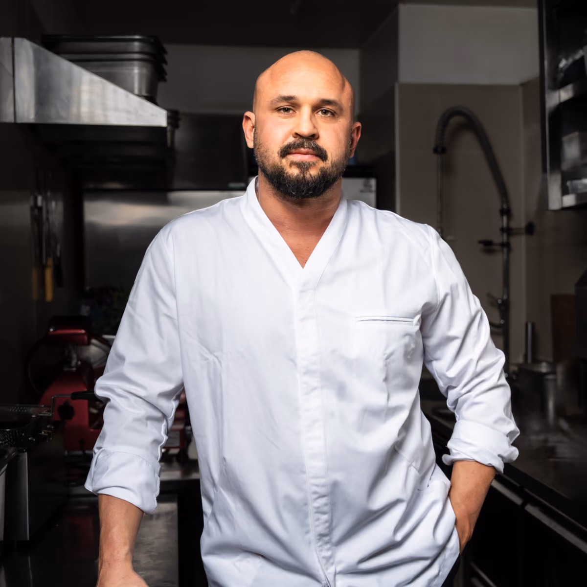 Chef with beard and bald head in a white chef's jacket stands in a professional kitchen.