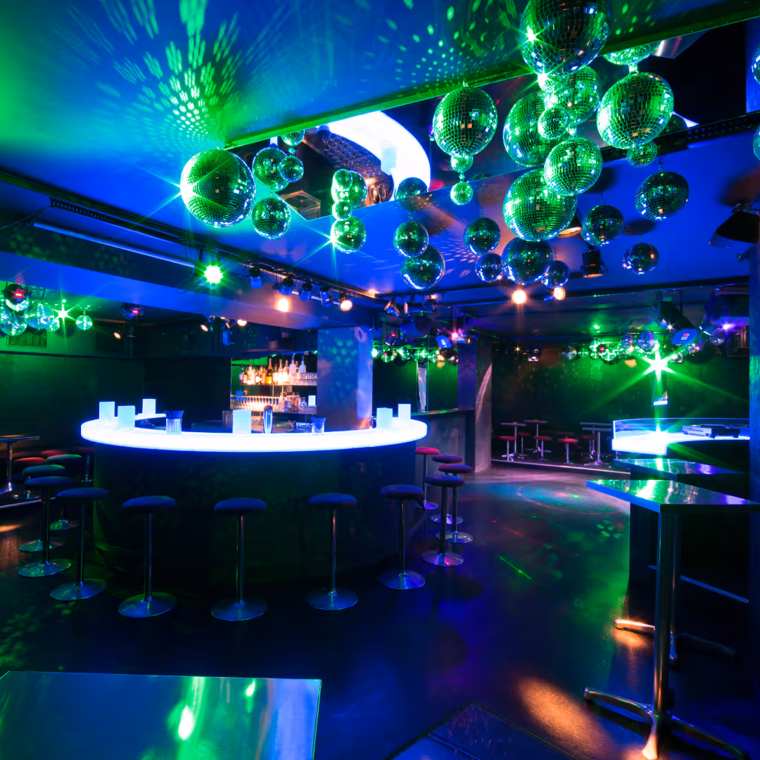 Innenansicht einer modernen Bar mit runder, leuchtender Theke, Barhockern und grünen, reflektierenden Disco-Kugeln an der Decke.