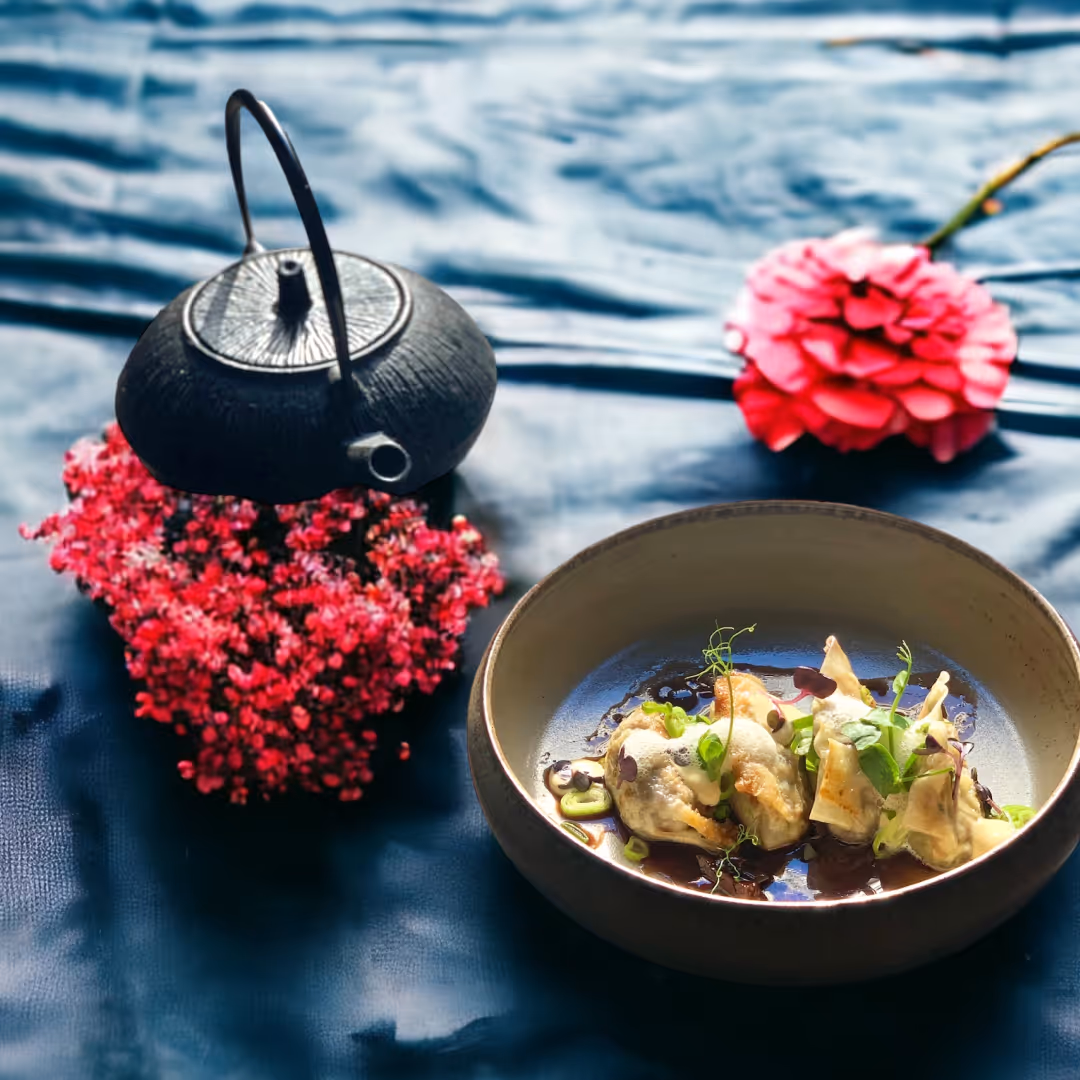 Schwarze Teekanne neben einer Schale mit dekorierten gedämpften Teigtaschen auf blauem Stoff mit roten Blumen.
