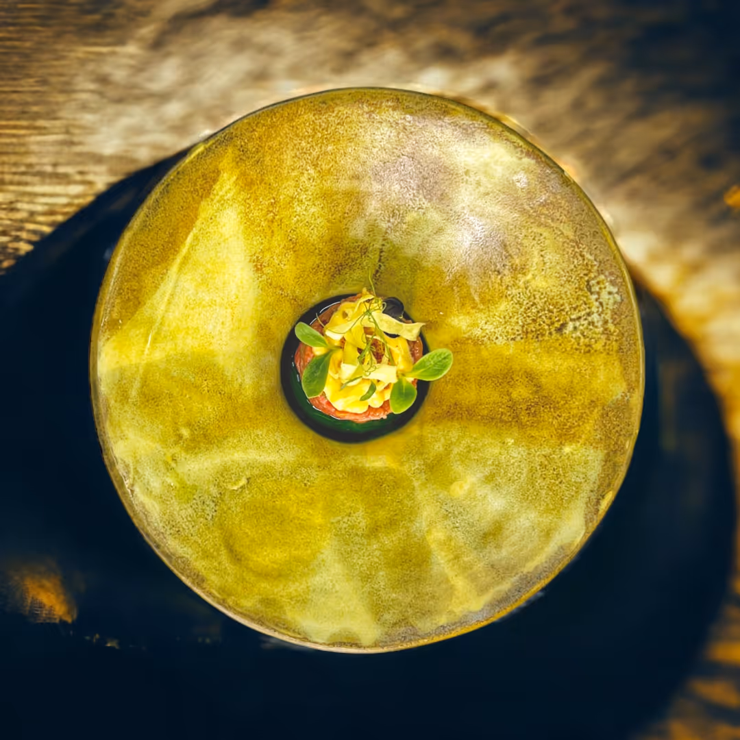 Fein angerichtetes Gericht mit gelben Blüten und grünen Blättern in der Mitte eines großen gelbgoldenen Tellers.