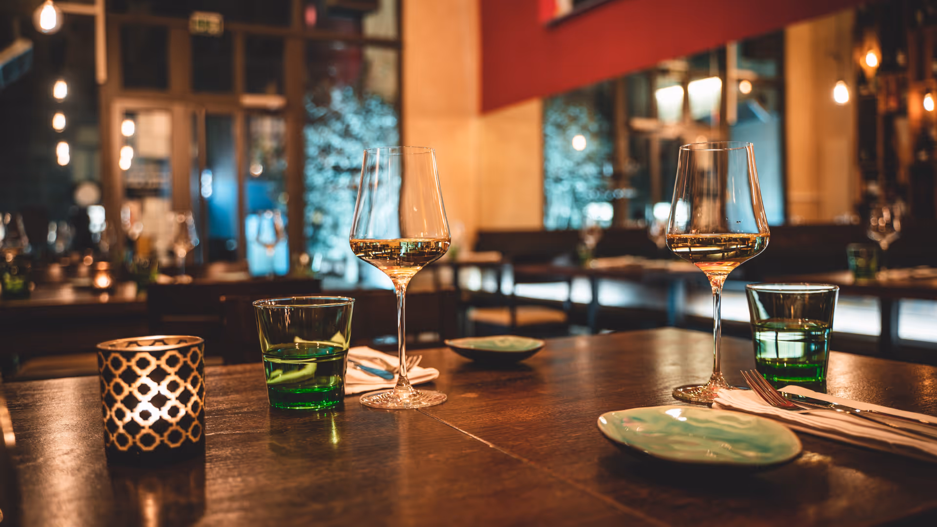 Ambiente in einem Restaurant mit zwei Weingläsern, grünen Wassergläsern, Tellern und einer Kerze auf einem Holztisch.