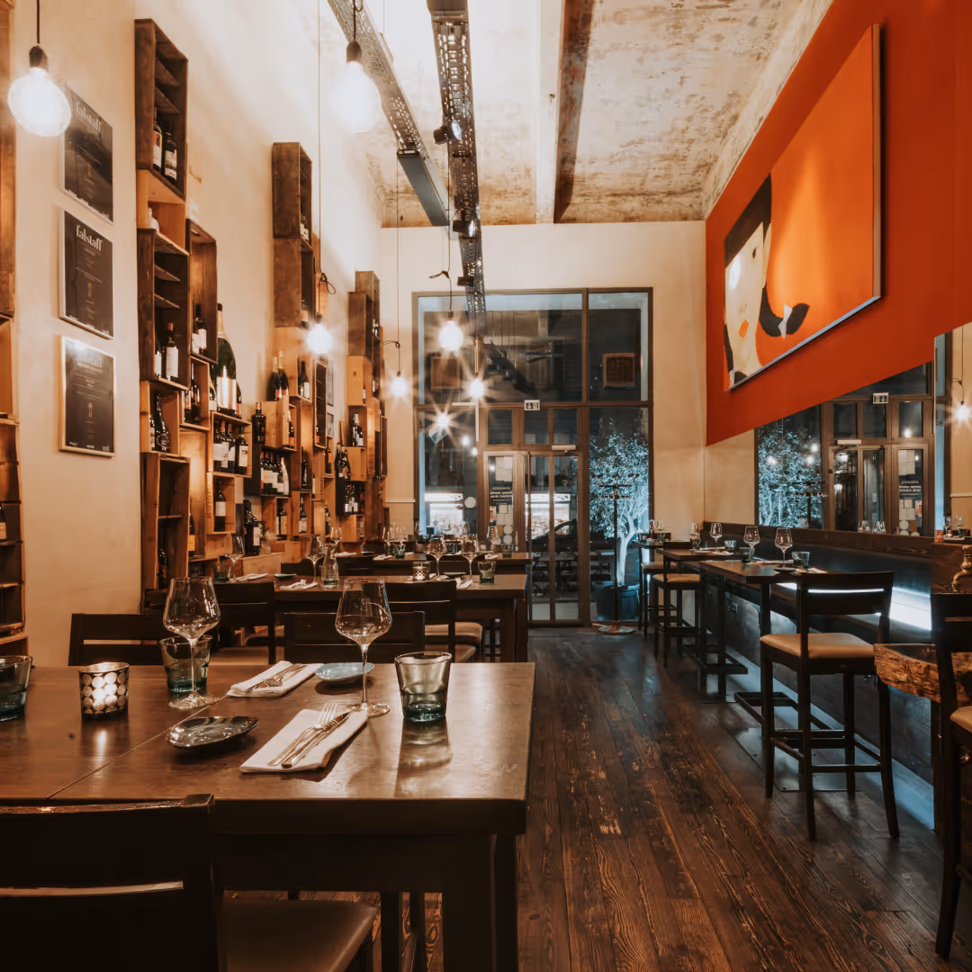 Gemütliches Restaurant mit Holzboden, Weinregalen an der Wand und modernem Kunstbild auf roter Wand.