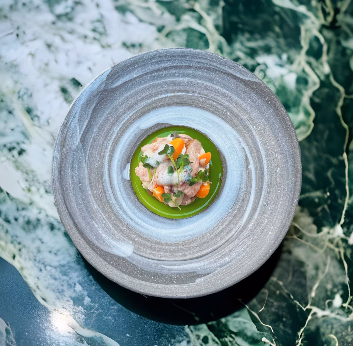 Gourmetgericht mit zartem Fleisch, kleinen Gemüsestücken und Mikrokräutern auf grünem Gelee, serviert auf einem grauen Teller mit weißen Pinselstrichen.