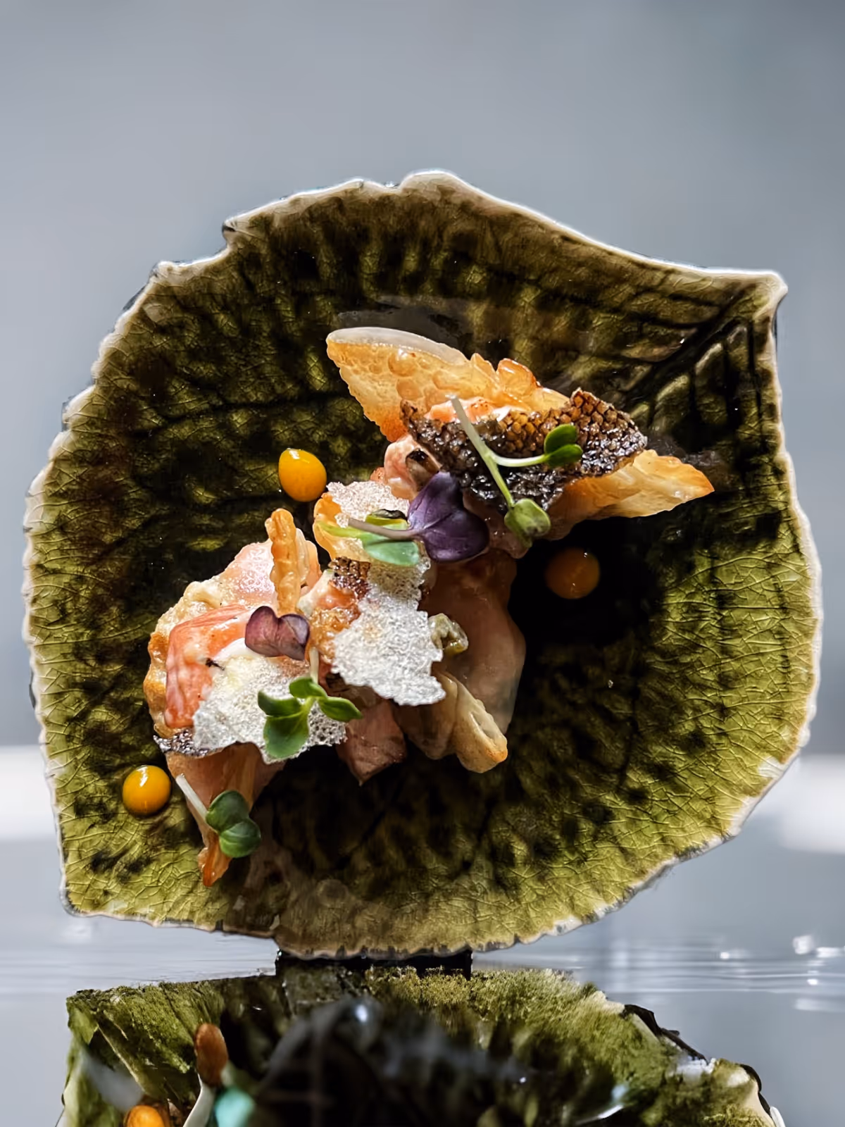 Kunstvoll angerichtetes Gericht mit Fischhaut, Garnelen und Mikrogrün auf einem grünen, blattähnlichen Teller.