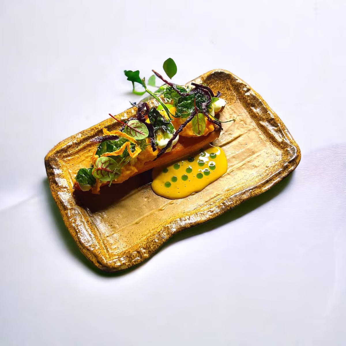 Kunstvoll angerichtetes Gericht mit orangefarbener Creme, grünen Blättern und gelber Sauce mit grünen Punkten auf rechteckigem braunem Teller.