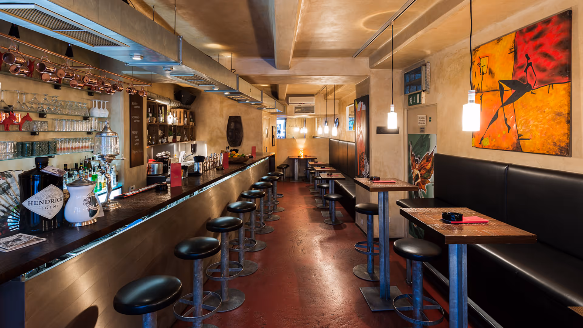 Innenraum einer gut beleuchteten Bar mit Hockern am langen Tresen und Tischen mit schwarzen Sitzbänken an der Wand.