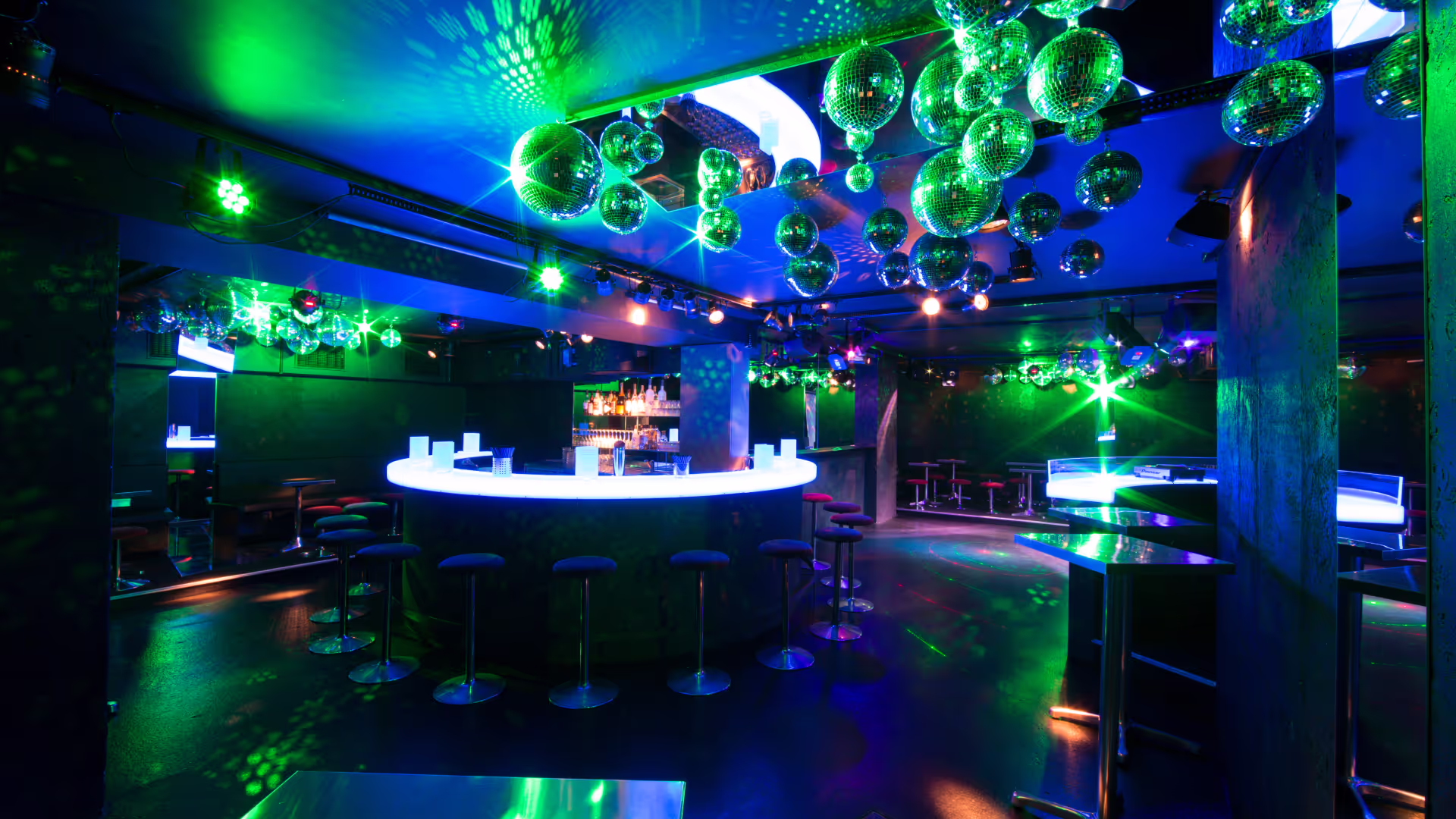 Dunkler Club mit rundem beleuchtetem Bartresen, Barhockern und zahlreichen Glas-Kugeln an der Decke bei grüner Beleuchtung.