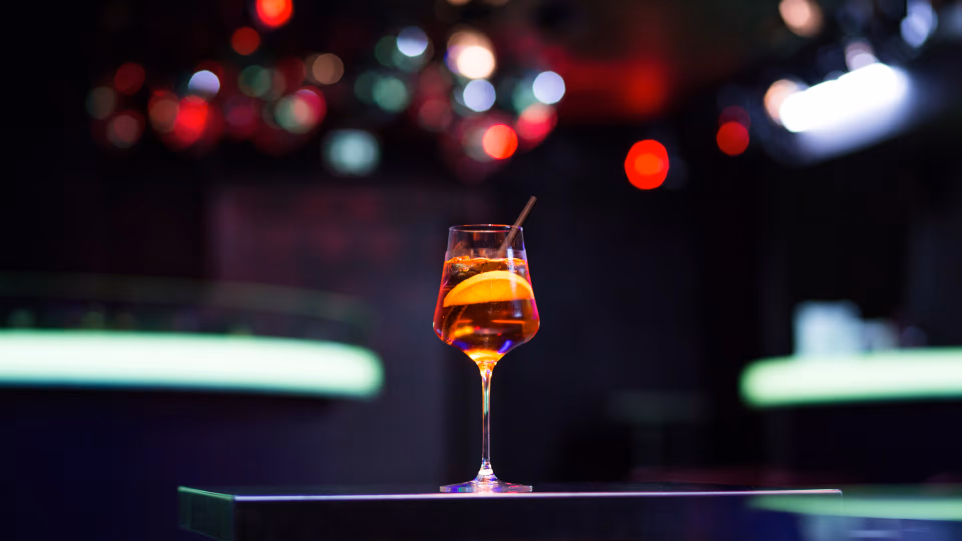 Ein Cocktailglas mit einem orangefarbenen Getränk, einer Orangenscheibe und einem Strohhalm vor unscharfem Bar-Hintergrund mit bunten Lichtern.