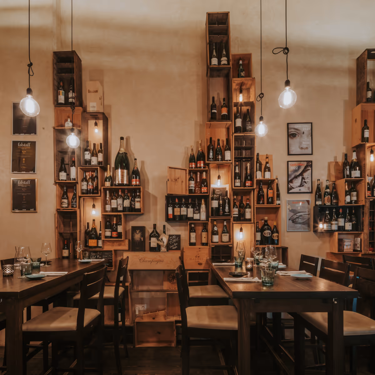 Gemütlicher Weinbar-Innenraum mit hölzernen Tischen, Stühlen, Weinflaschen in Holzkästen an der Wand und hängenden Glühbirnen.