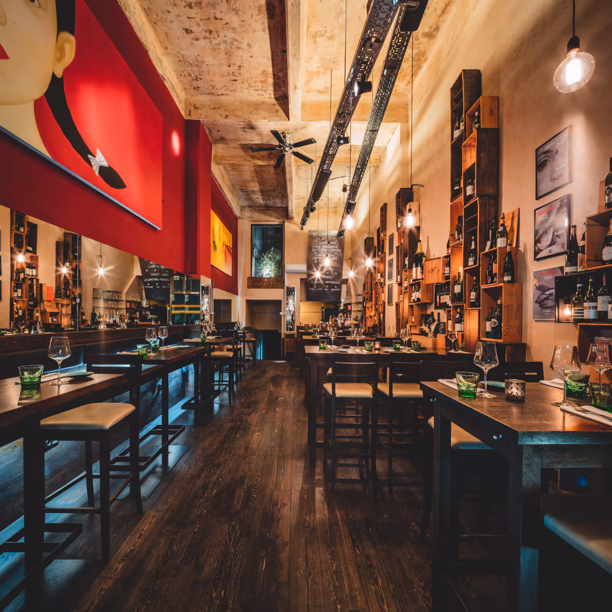 Modernes Restaurant mit dunklem Holzboden, hölzernen Tischen und Stühlen, roten und beigen Wänden, Weinregalen und stimmungsvoller Beleuchtung.