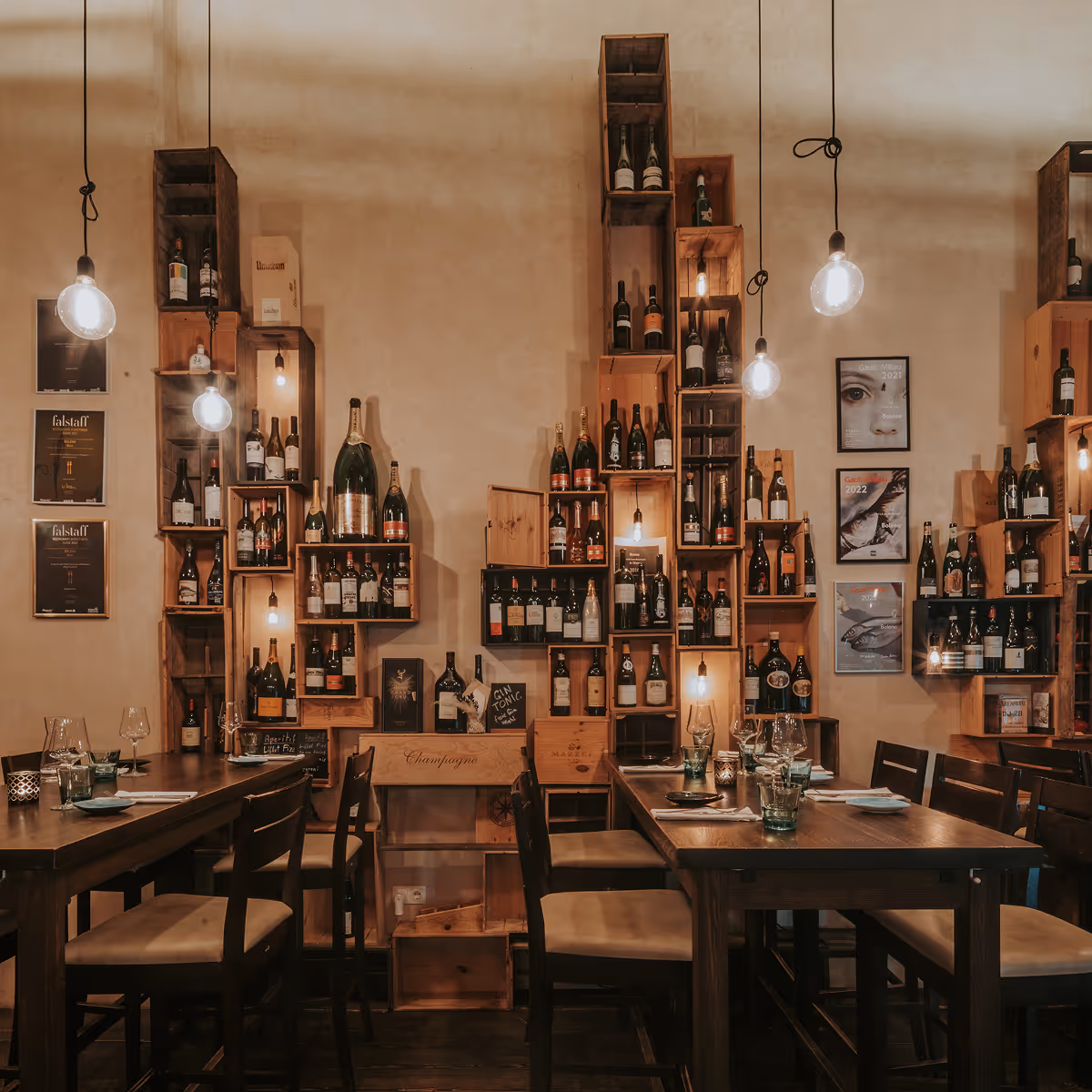 Gemütlicher Gastraum mit Holztischen, Stühlen und einer Wand mit hängenden Holzregalen, die Weinflaschen und dekorative Lichter präsentieren.