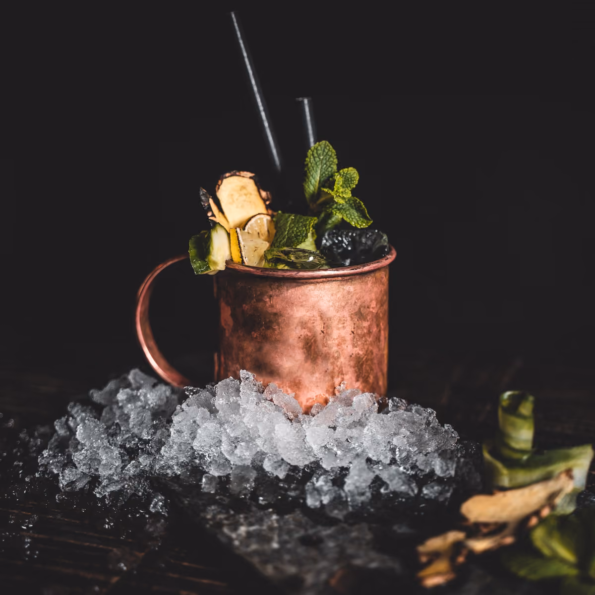 Kupferbecher mit Eis, Gurke, Ingwer, Limette und Minze vor dunklem Hintergrund.