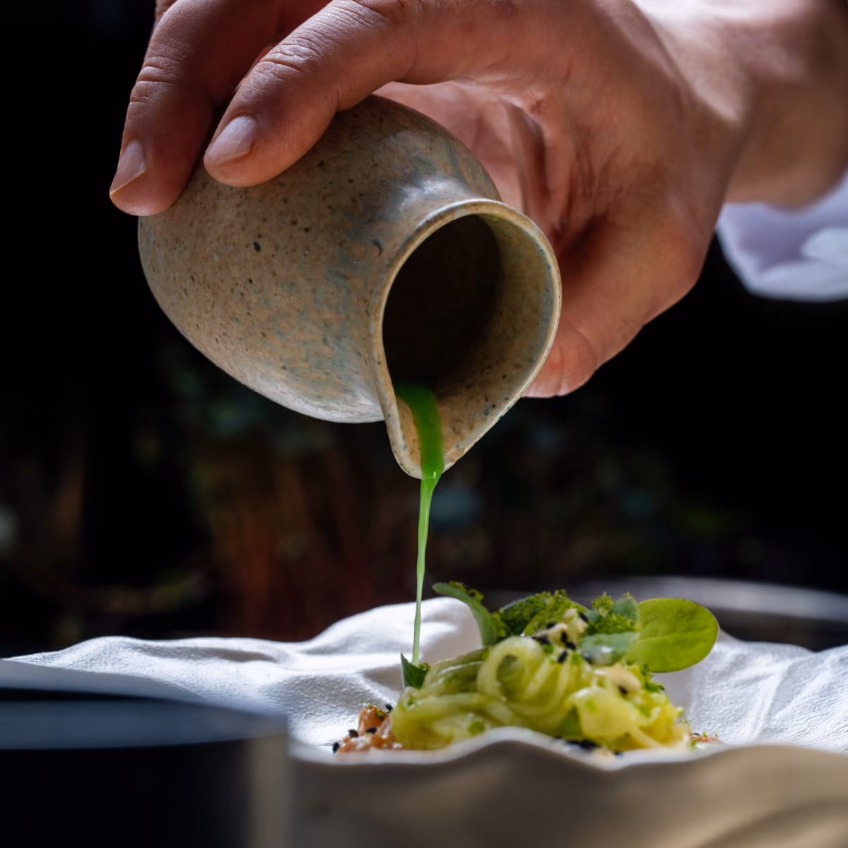 Hand Pours Green Dressing from Clay Jug Onto Salad with Cucumbers and Herbs.