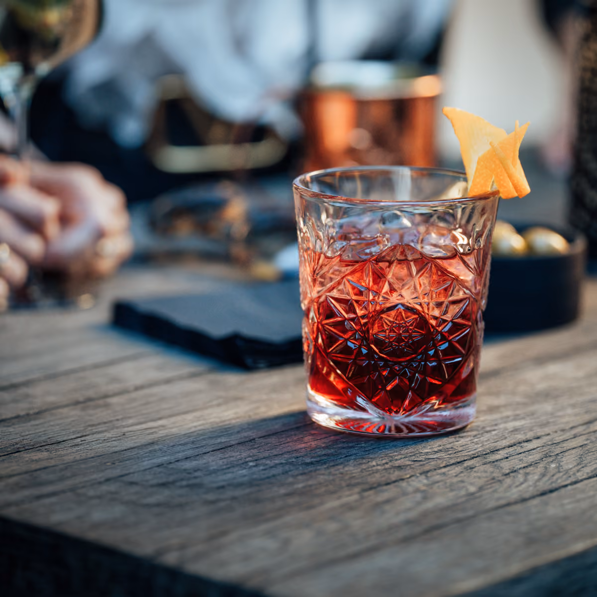 Verzierter Glas mit rotem Cocktail, Eiswürfeln und Orangenschale auf Holztisch.