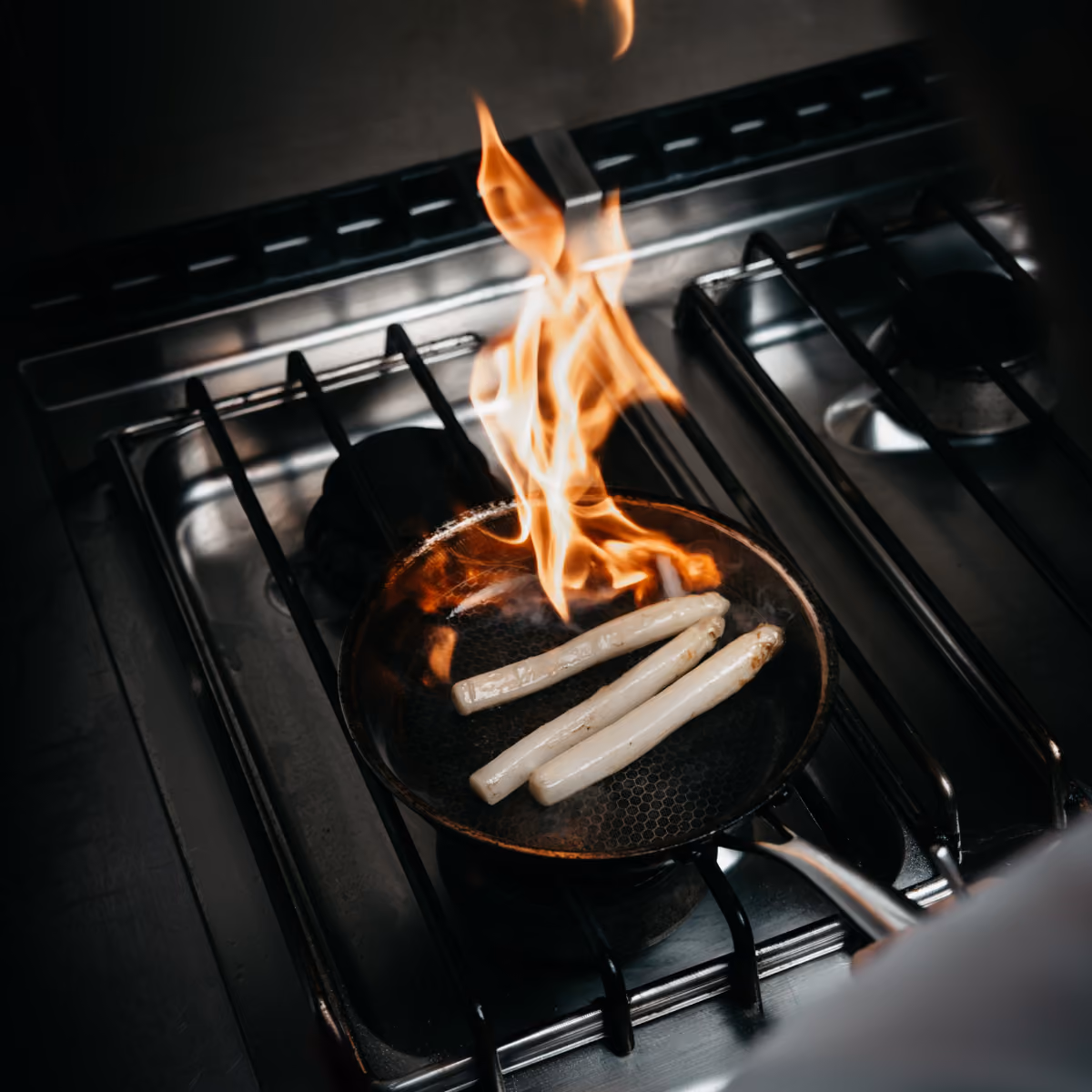 Drei weiße Spargelstangen braten in einer Pfanne über Flammen auf einem Gasherd.