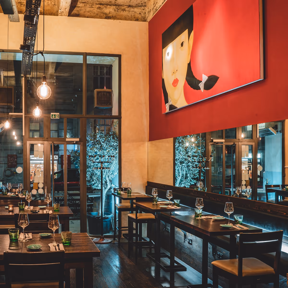 Gemütliches Restaurant mit Holztischen, gedeckten Gläsern und einem großen Gemälde eines Mädchens mit Zöpfen an roter Wand.