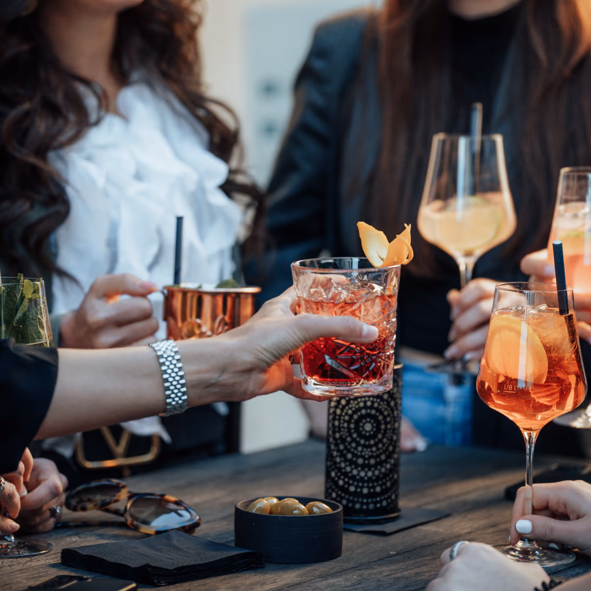 Menschen stoßen bei einem Treffen mit verschiedenfarbigen Cocktails und Snacks an einem Holztisch an.