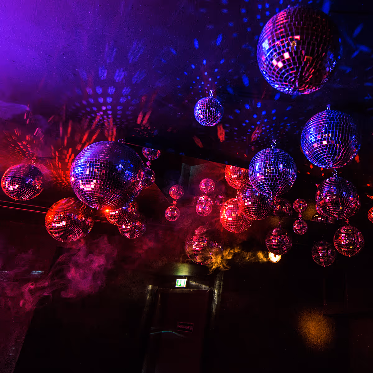 Mehrere Disco-Kugeln hängen von der Decke und reflektieren rote und blaue Lichter in einem dunklen Raum.