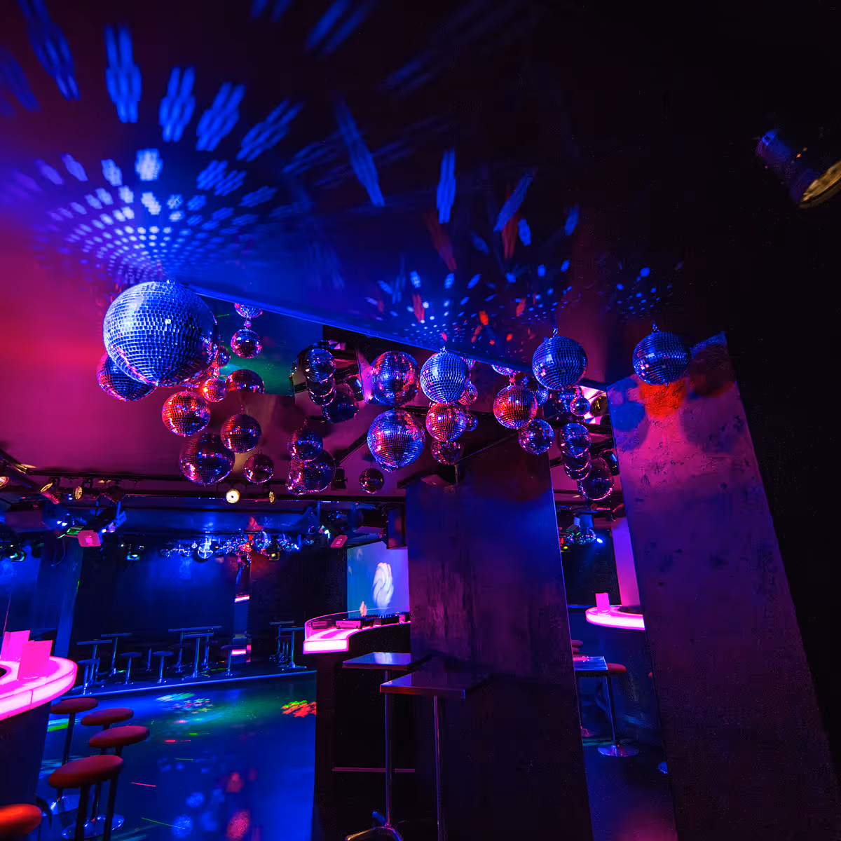 Innenraum einer Disco mit mehreren Spiegelkugeln an der Decke, neonblauem und rosafarbenem Licht und Barhockern.