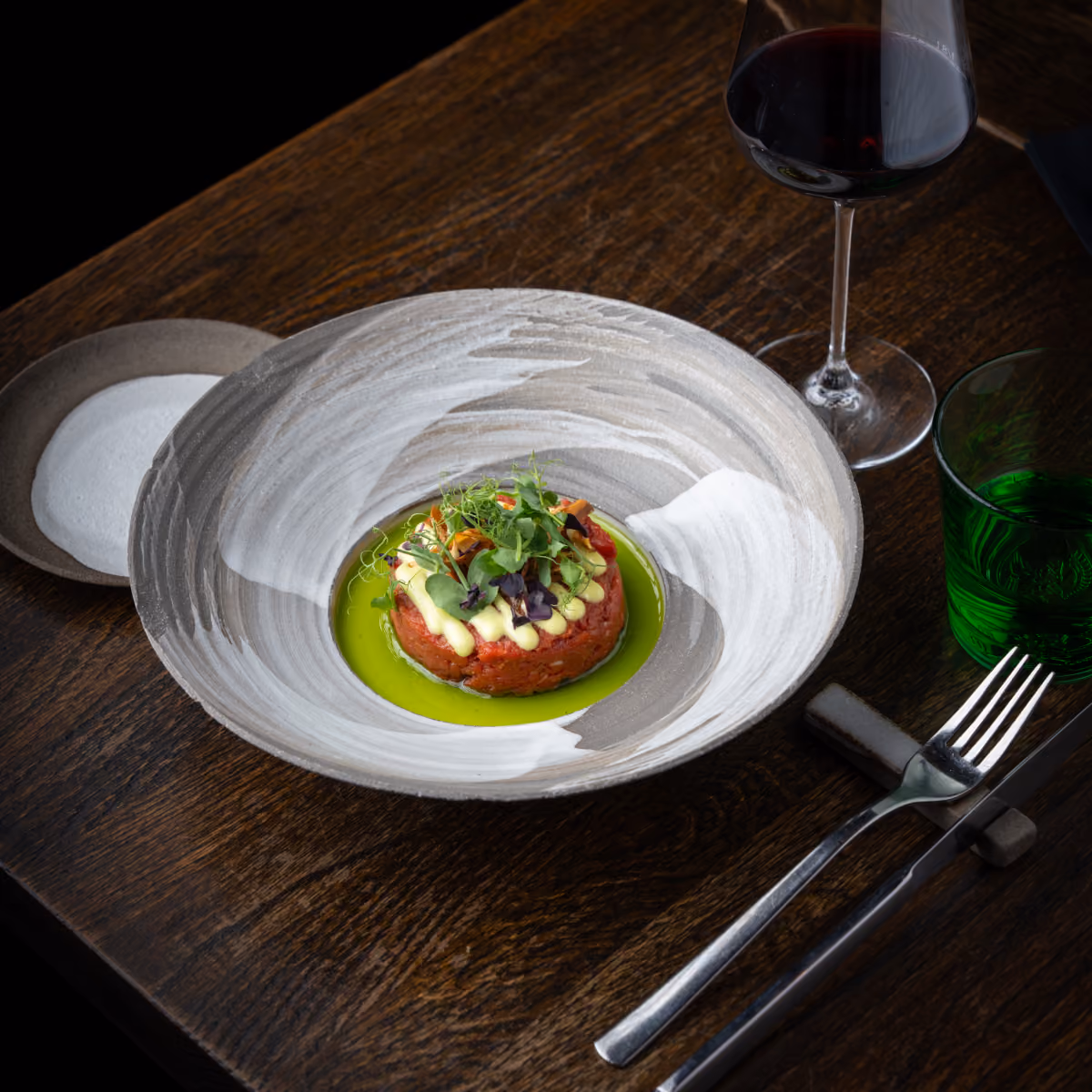 Plate with beef tartare, green oil, sauce, herbs, next to wine glass, green glass, fork and knife.