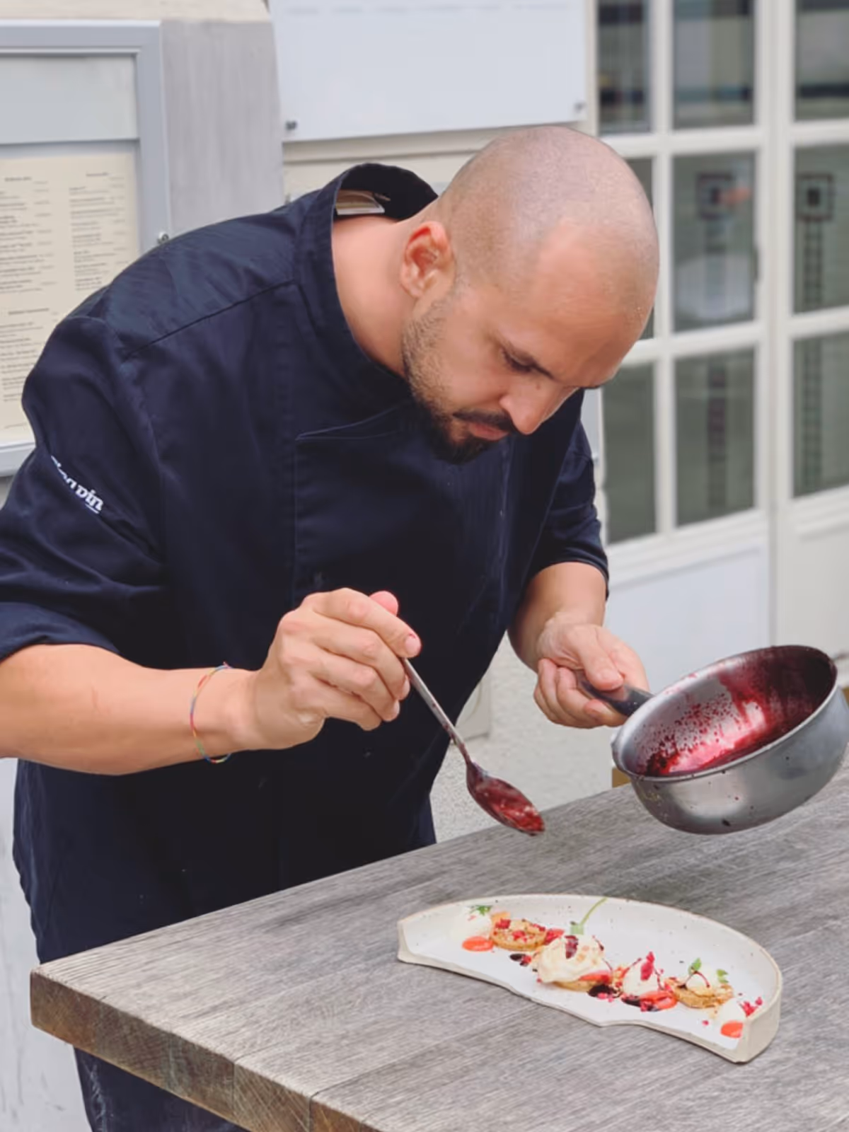 Koch trägt schwarze Jacke und garniert mit einem Löffel eine künstlerisch angerichtete Nachspeise auf einem weißen Teller.