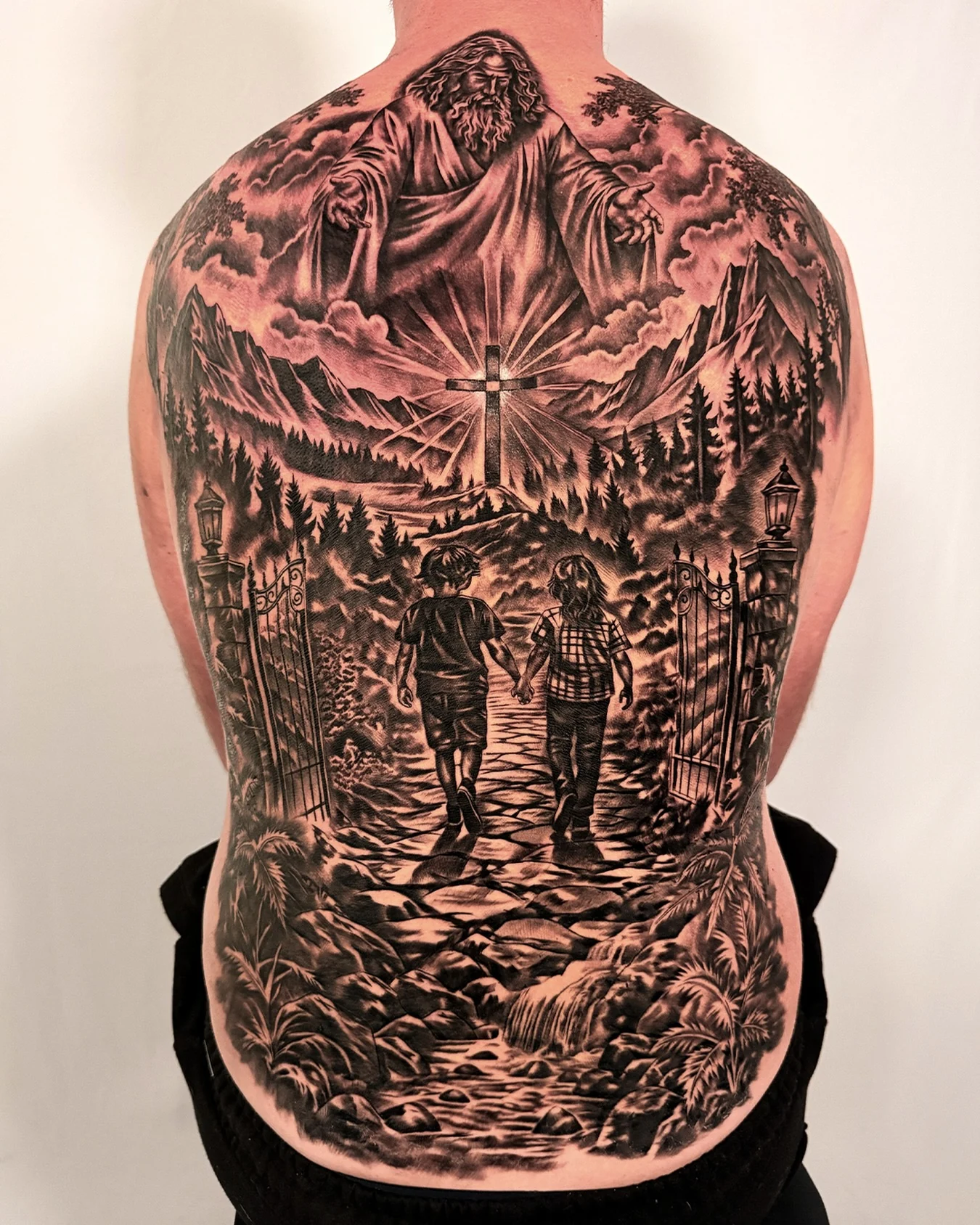 Black and grey full-back tattoo with a robed religious figure, central cross, mountains and two children holding hands, by Joshua Hancox