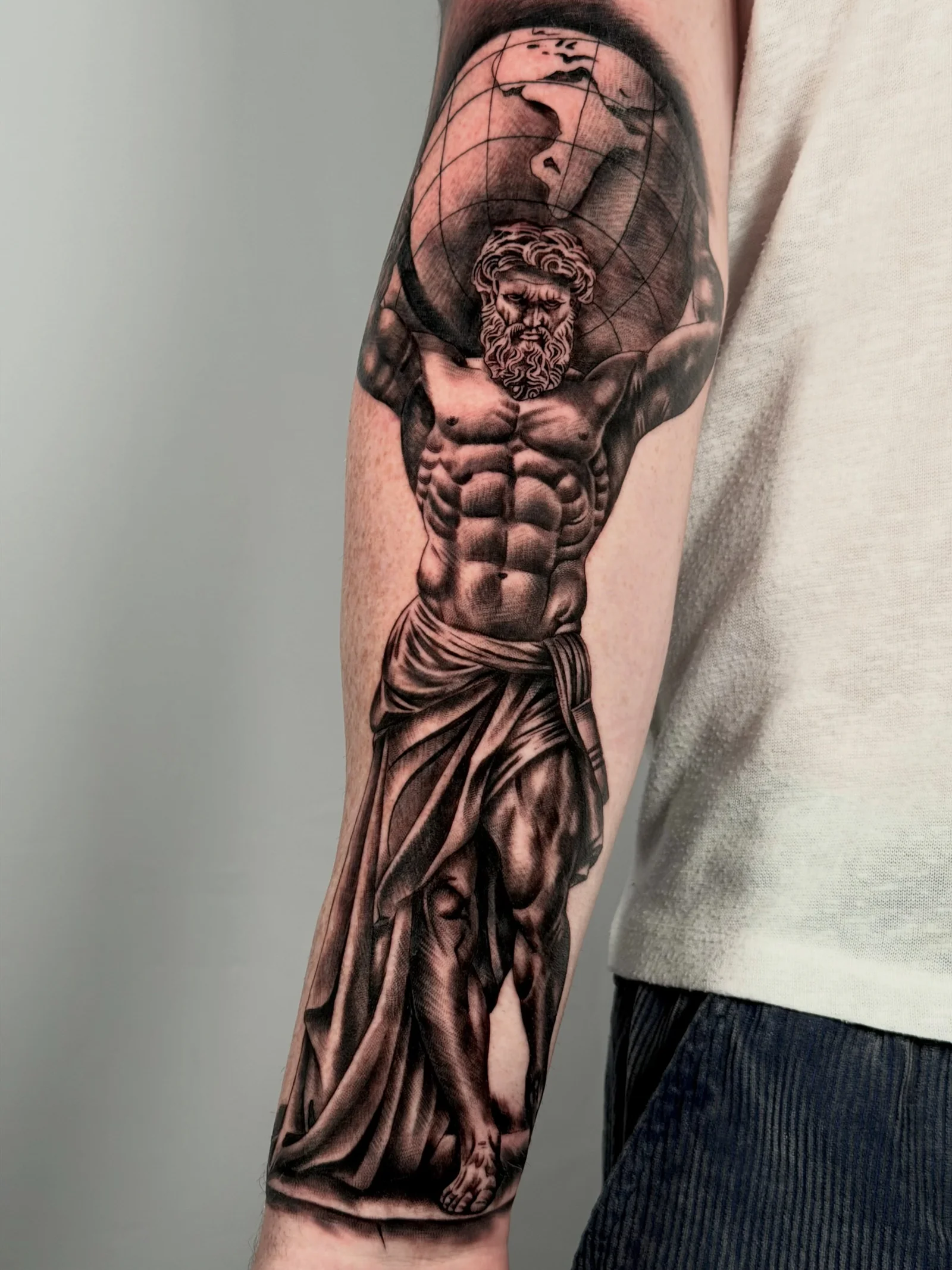 Black and grey forearm tattoo of Atlas carrying a globe, with sculptural shading and draped fabric, by Joshua Hancox