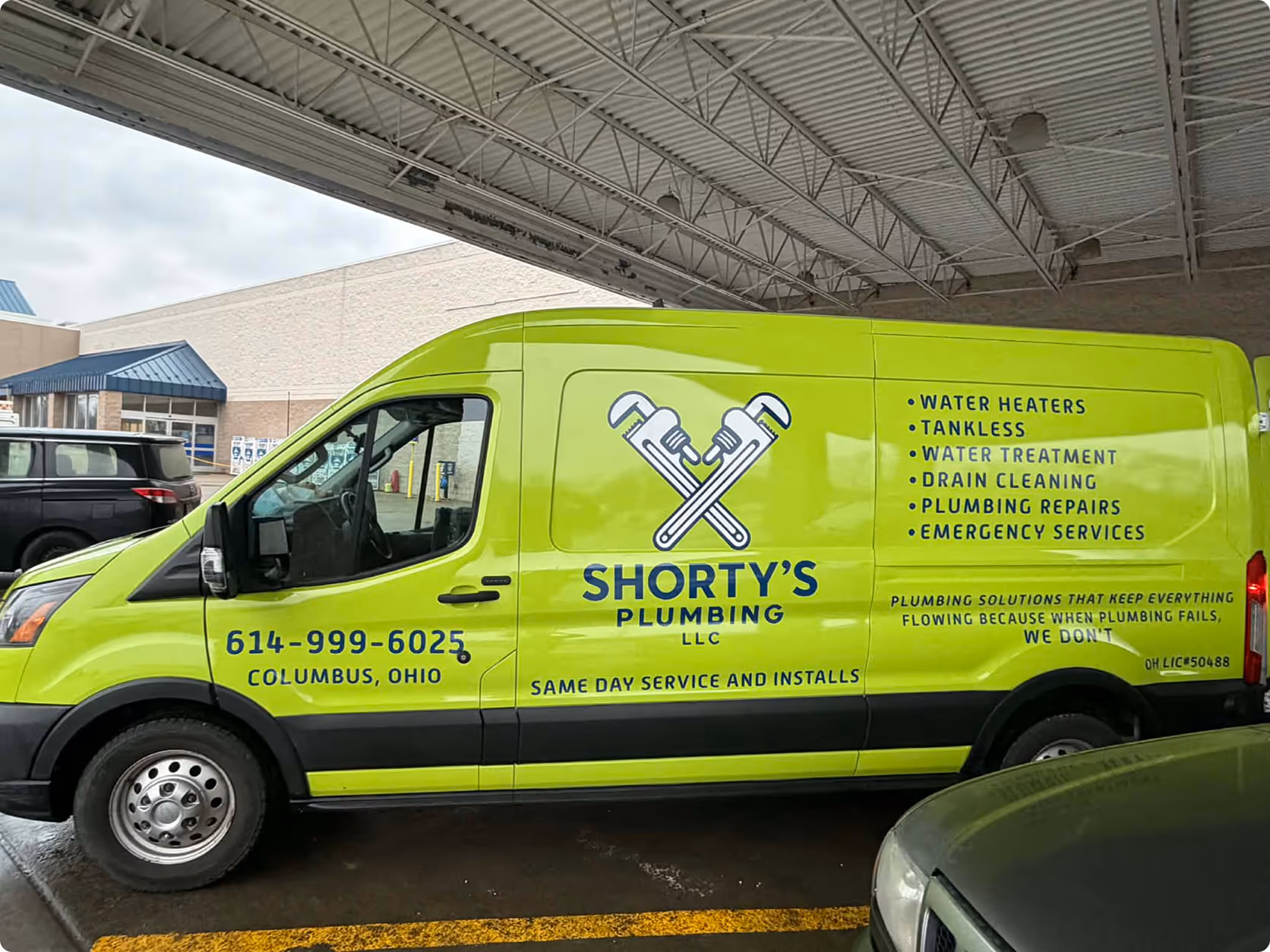Bright green Shorty's Plumbing van with contact number, services offered, and company slogan parked under a metal canopy.