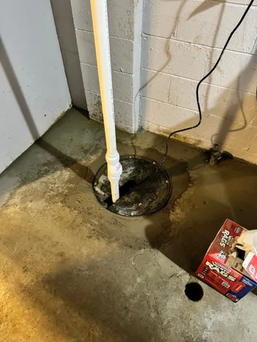 Basement corner with a sump pump installed in the floor, white PVC pipe, and a box of tools nearby.