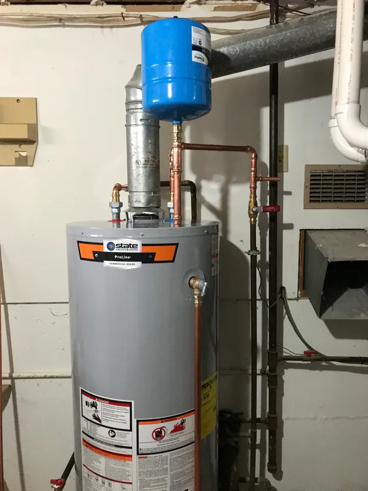 Gray State Water Heaters ProLine commercial-grade water heater with a blue expansion tank attached on top, set against a basement wall with exposed pipes and vents.