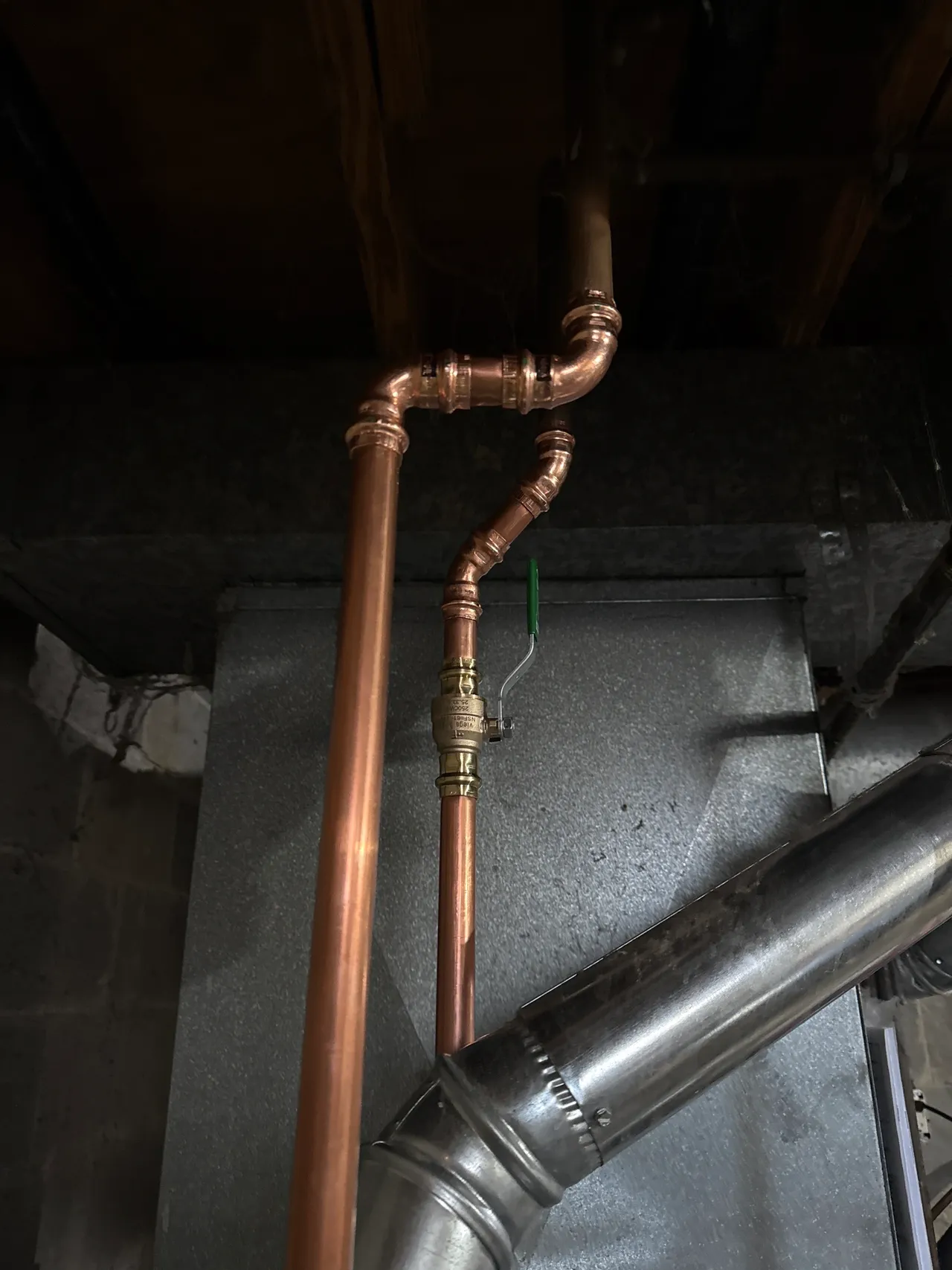 Copper plumbing pipes with a green-handled valve connected near a metallic duct in a dimly lit basement ceiling.