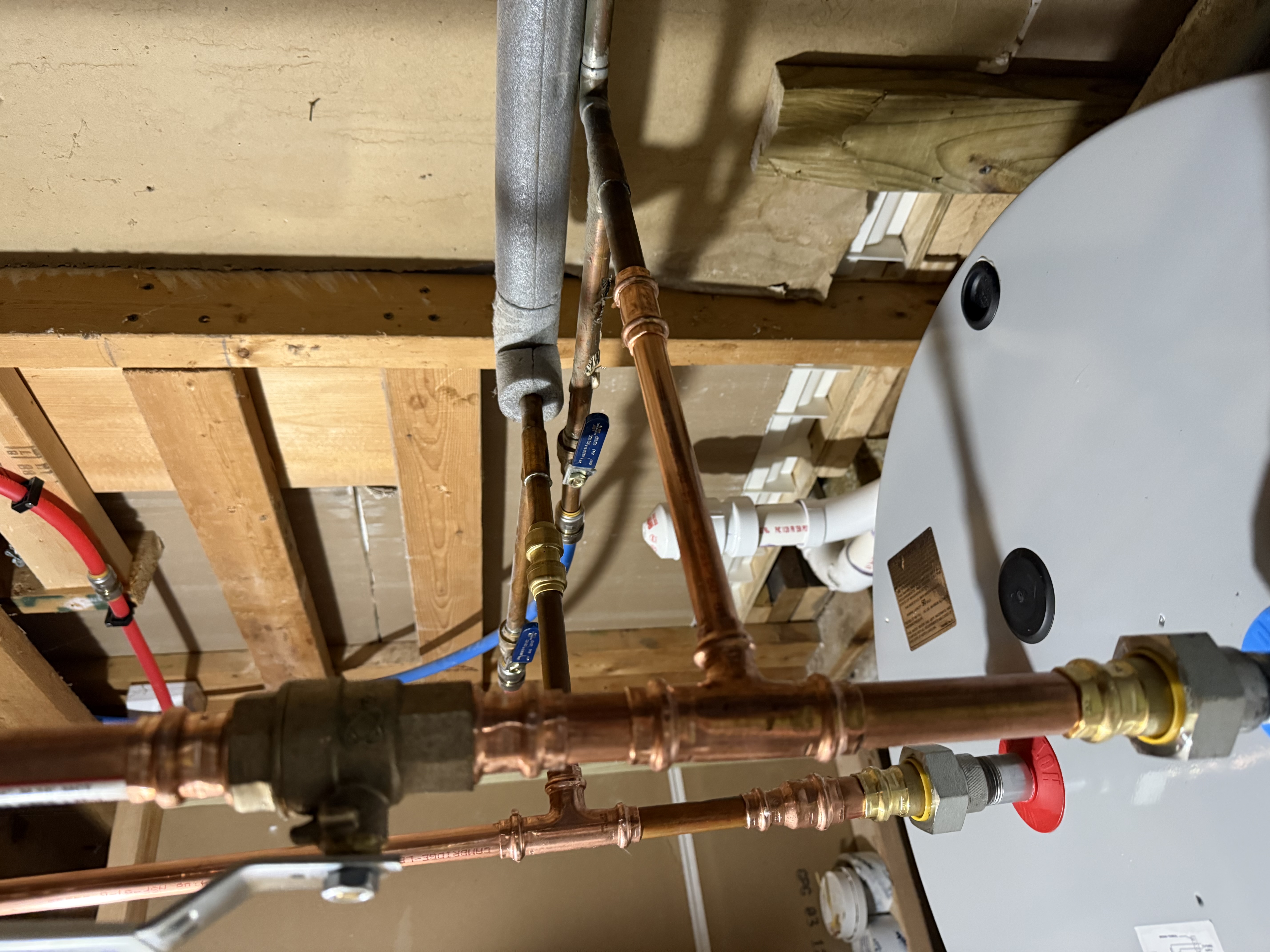 Copper water pipes connected to a white water heater tank with wooden studs and insulation in the background.