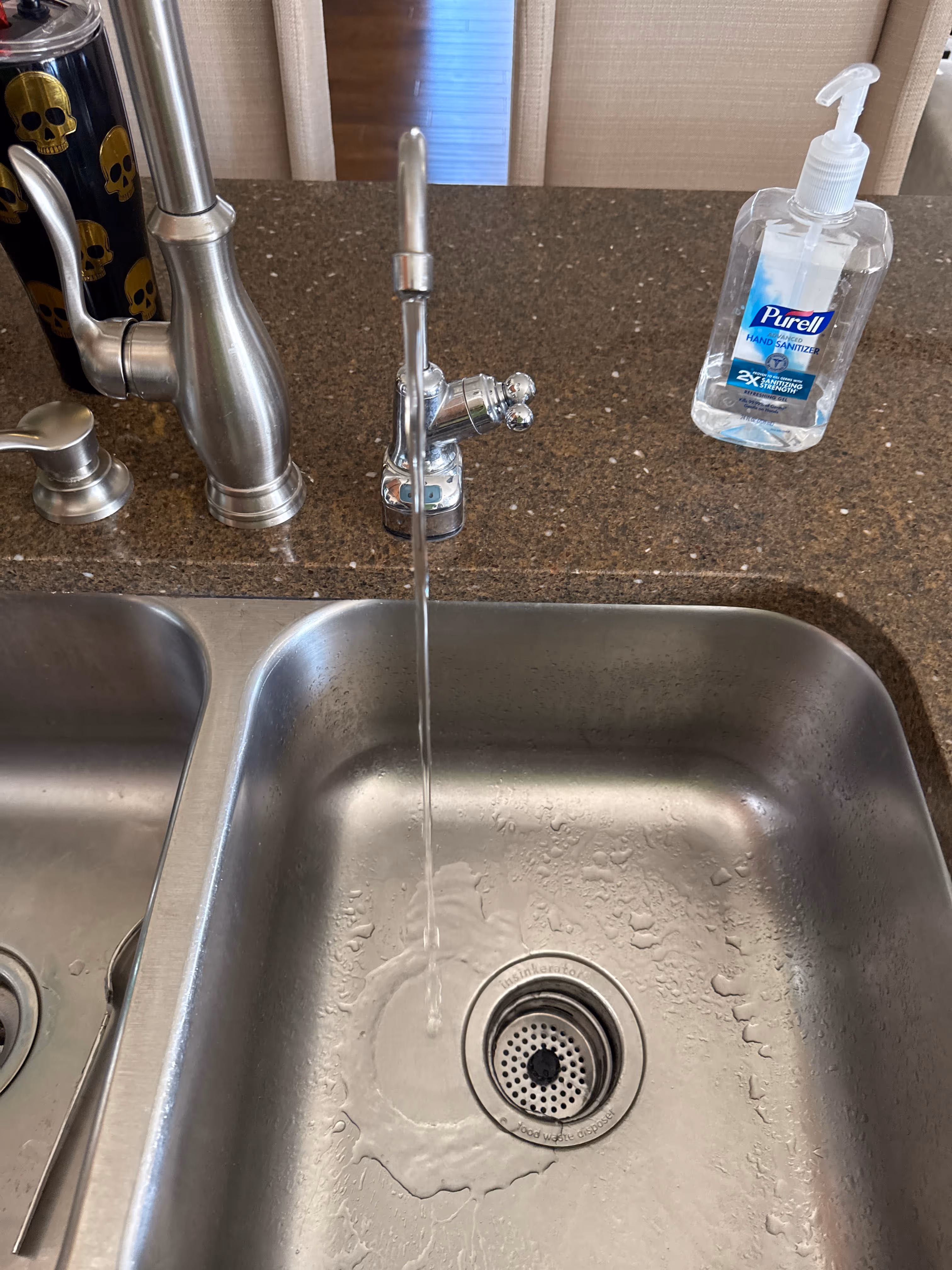 Stainless steel kitchen sink with running water from a faucet and a bottle of Purell hand sanitizer on the brown countertop.