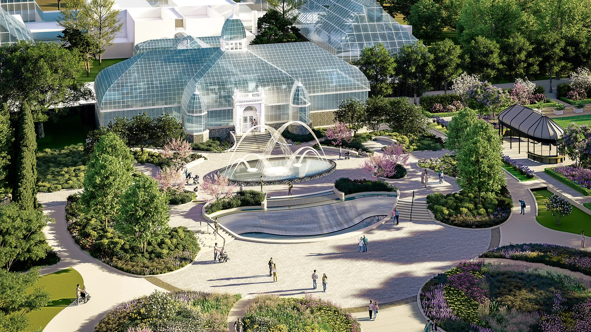 Aerial view of a glass conservatory surrounded by green trees, colorful flower beds, paved walkways, a water fountain, and people walking and sitting in a landscaped park.