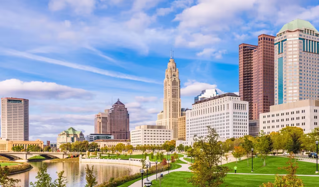 View of Columbus, Ohio skyline with the Scioto Mile waterfront park in the foreground under a partly cloudy sky.