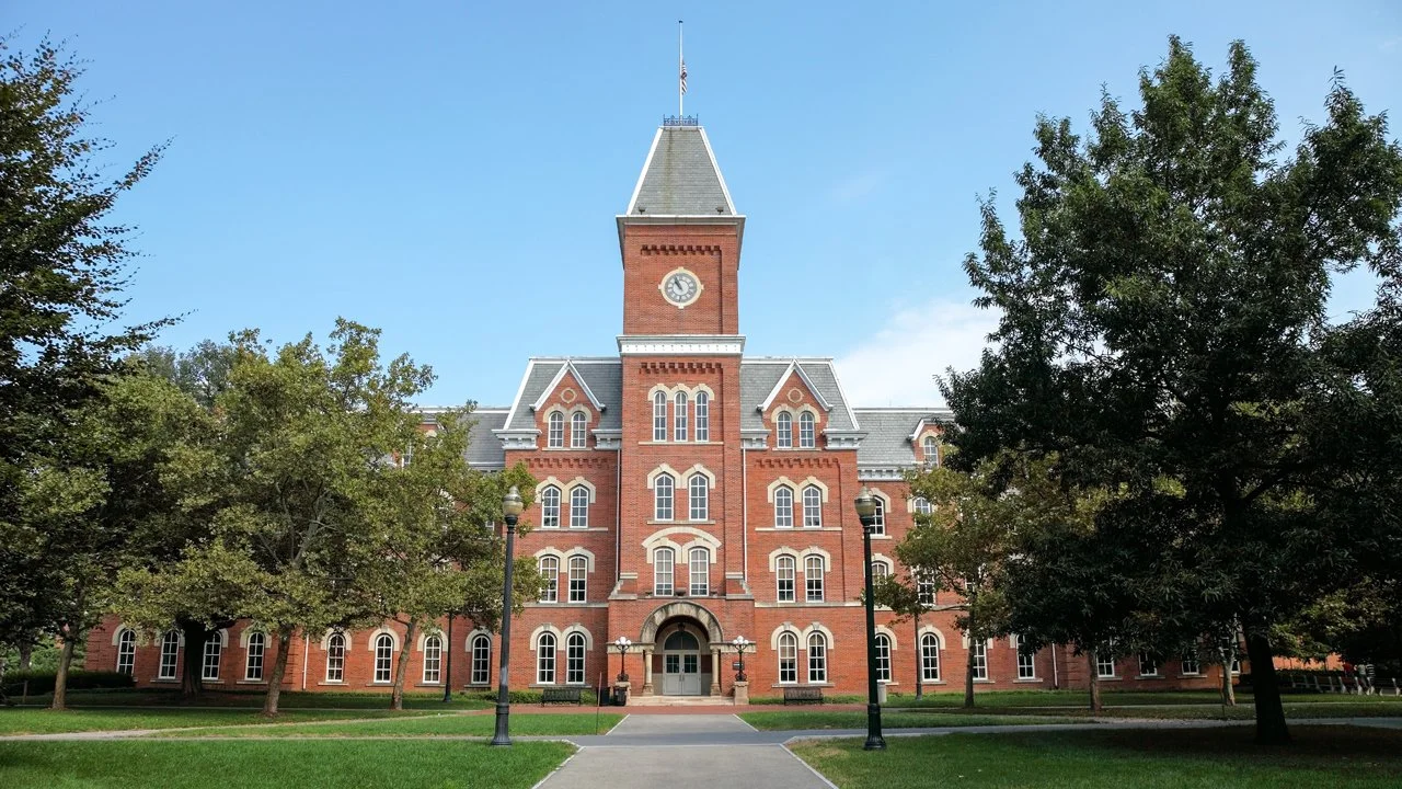 Historic red brick university building with a tall clock tower and surrounding green trees under a clear blue sky.