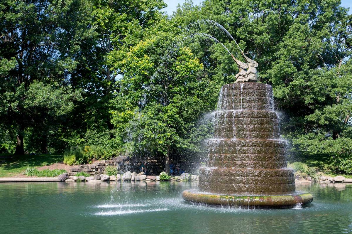 Tiered stone fountain in a pond with sculptures of two children at the top, spraying water, surrounded by lush green trees.